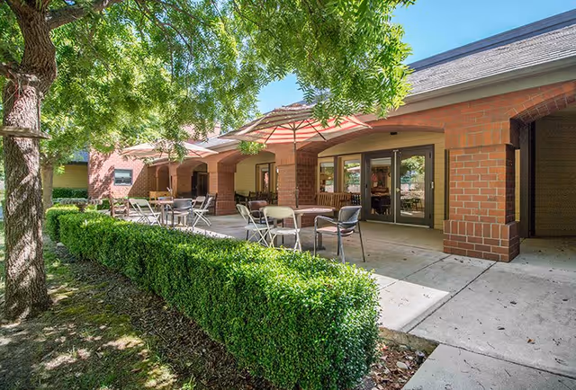 Outdoor patio area at Summerfield of Roseville with tables, chairs, and umbrellas set along a walkway next to a building with brick pillars and large windows, surrounded by green hedges and trees.