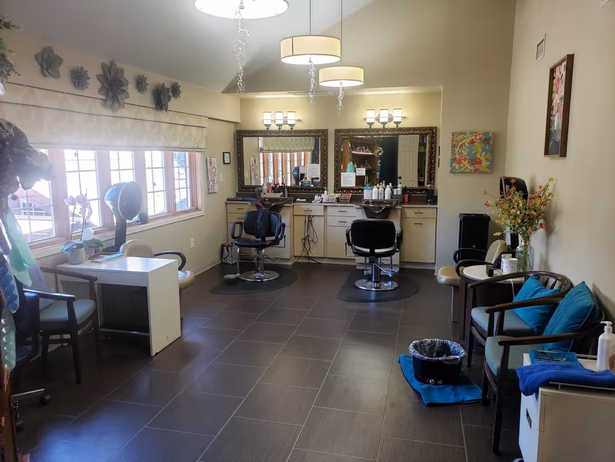 Bright salon-style room with hair-styling chairs, mirrors, seating, and decorative accents in a senior living facility.