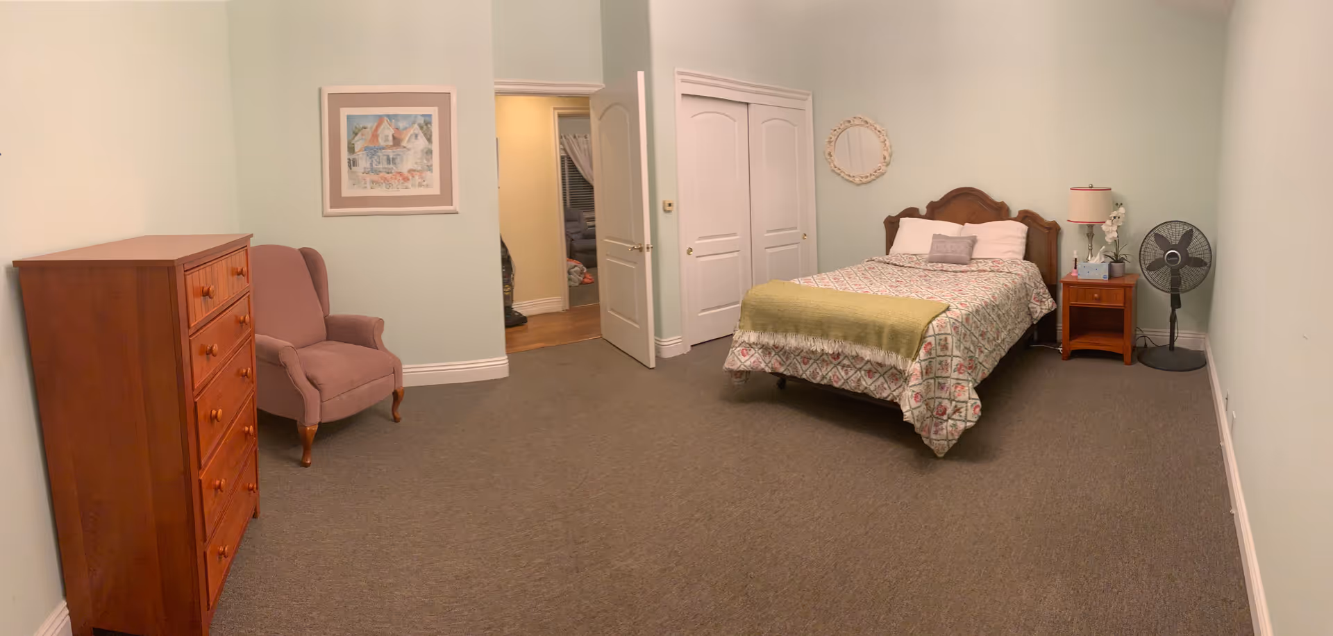 A bedroom with a bed covered in a patterned quilt and a green throw blanket. There is a wooden nightstand with a lamp, tissue box, and a small plant next to the bed. A standing fan is positioned near the nightstand. On the opposite side of the room, there is a wooden dresser and a purple upholstered armchair. The walls are painted light green, and a framed picture hangs above the armchair. The room has a carpeted floor and a closet with white double doors. An open door leads to another room with hardwood flooring.