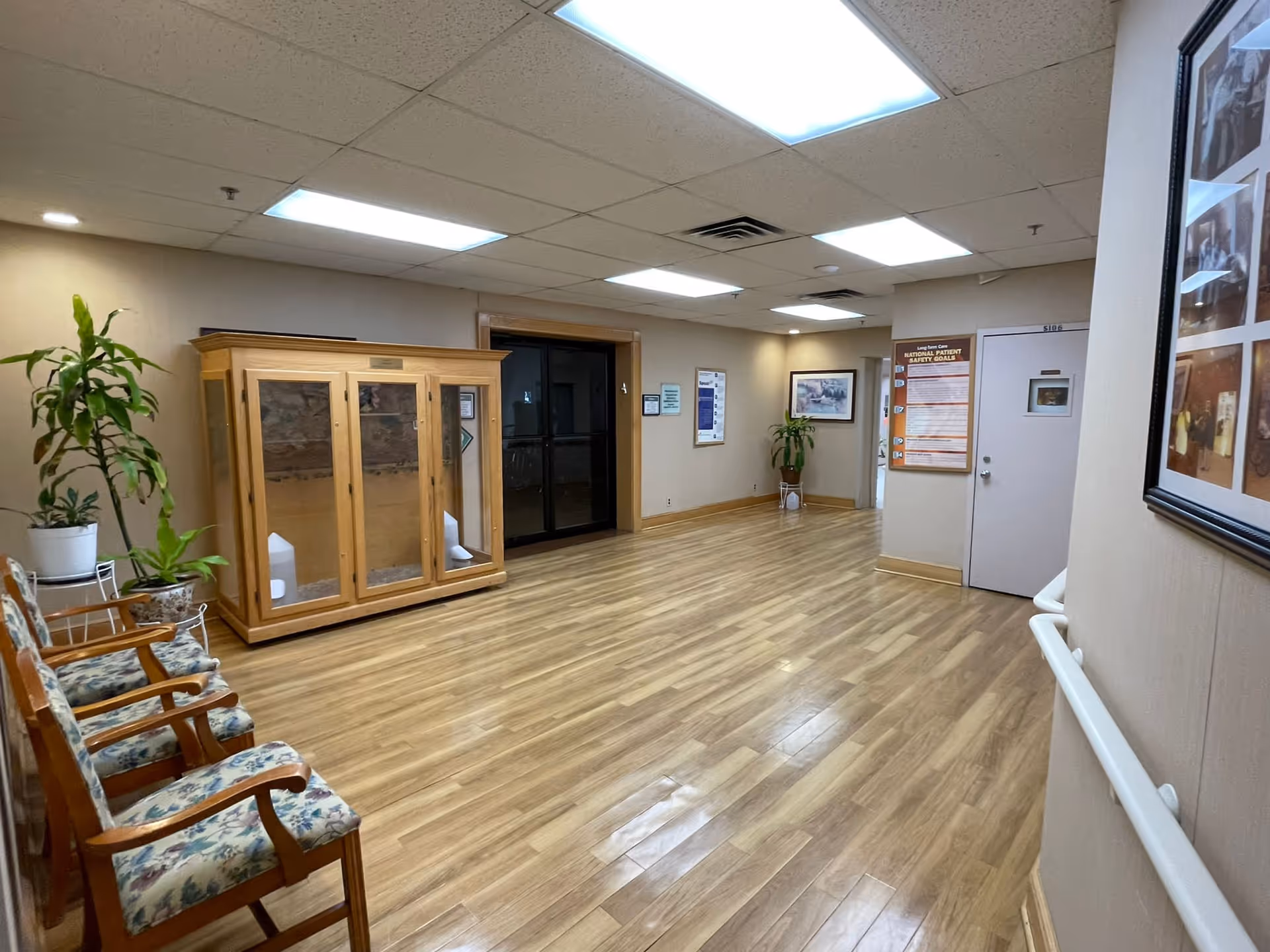A clean, well-lit hallway in a senior living facility with wooden flooring, floral cushioned chairs along the left wall, potted plants, framed pictures on the walls, and a glass display cabinet. The ceiling has fluorescent lights, and there is a white door with a safety poster next to it on the right side.