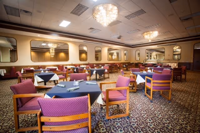 A spacious dining room with multiple tables covered in dark blue tablecloths and white napkins, surrounded by wooden chairs with purple cushions. The room features patterned carpet flooring, large mirrors on the walls, and elegant chandeliers hanging from the ceiling.