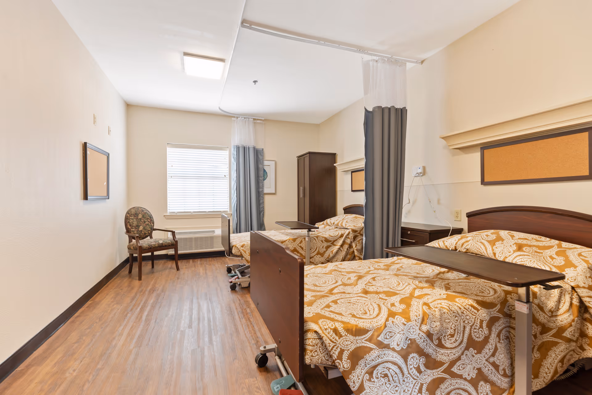 A senior living facility bedroom with two hospital-style beds, each covered with a patterned beige and white bedspread. Each bed has an overbed table and a privacy curtain that can be drawn between them. The room has wood flooring, a window with blinds, a floral upholstered chair, a wooden cabinet, and two cork bulletin boards on the walls.