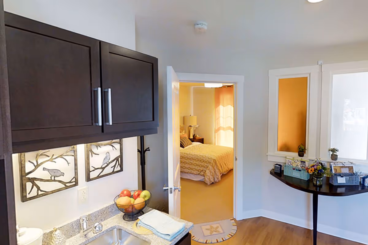 Interior view of a senior living facility showing a kitchenette area with dark wood cabinets, a sink, and a bowl of fruit on the counter. Through an open door, a bedroom with a bed covered in a yellow patterned bedspread is visible. To the right, there is a small table with decorative items and plants, and two windows with orange and white shades.