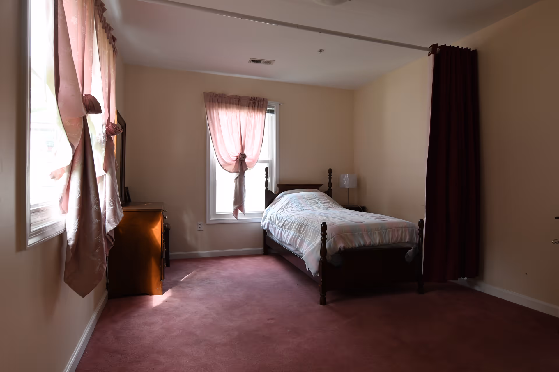 Single-occupancy bedroom with a wooden bed, pink curtains, a dresser, and mauve carpet.