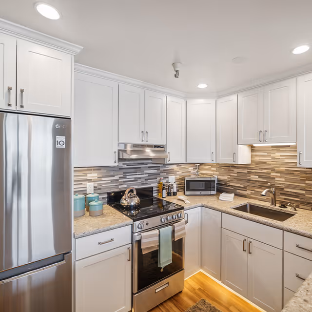 Modern kitchen with white cabinets, stainless steel refrigerator and stove, a microwave on the countertop, a sink with a faucet, and a backsplash with horizontal striped tiles in neutral tones.