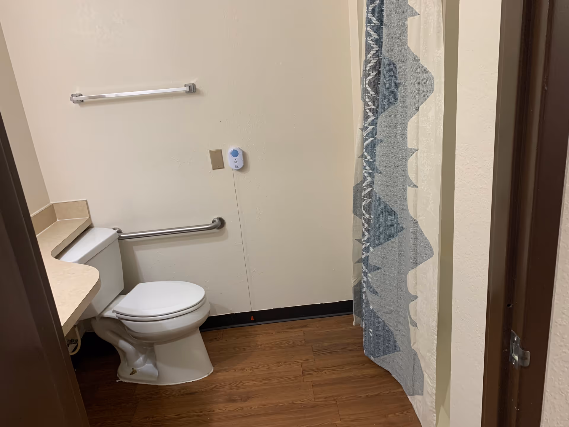 A bathroom with a white toilet, a beige countertop, a silver grab bar on the wall, a towel rack, and a shower curtain with a blue and white geometric pattern. The floor is wood-style laminate.