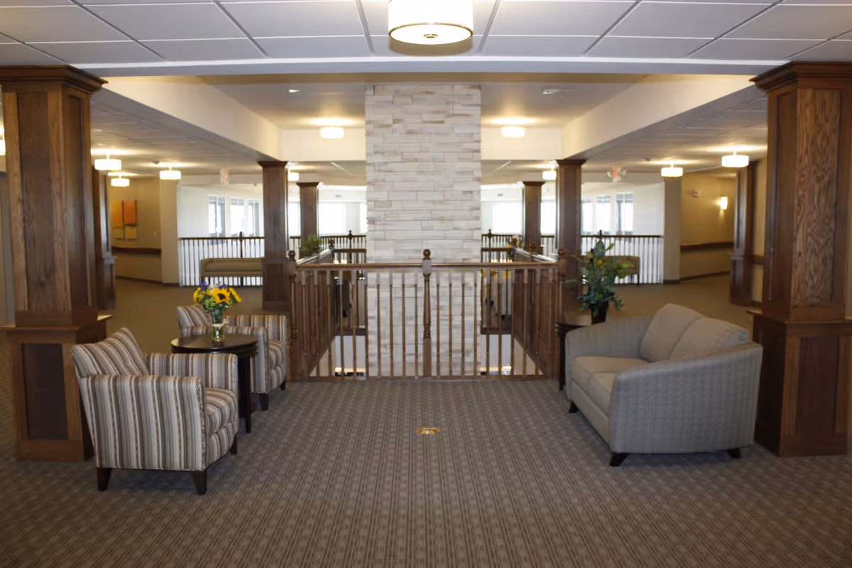 A spacious upstairs common area with upholstered chairs and a sofa arranged around a central railing and stone-clad column, flanked by wooden pillars and overhead lights.