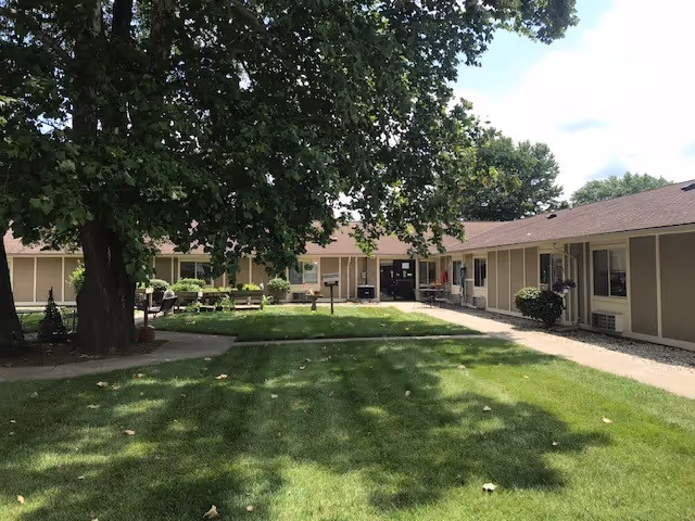 Outdoor courtyard area of Swiss Villa Nursing & Rehabilitation featuring a large tree providing shade, green grass, paved walkways, and a single-story building surrounding the courtyard with windows and doors.