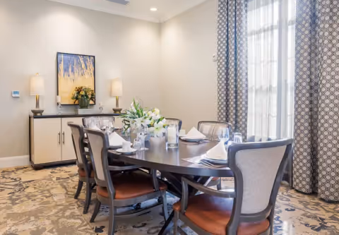 Formal dining room with an oval table set for six, upholstered chairs, a sideboard with lamps and artwork, and patterned curtains.