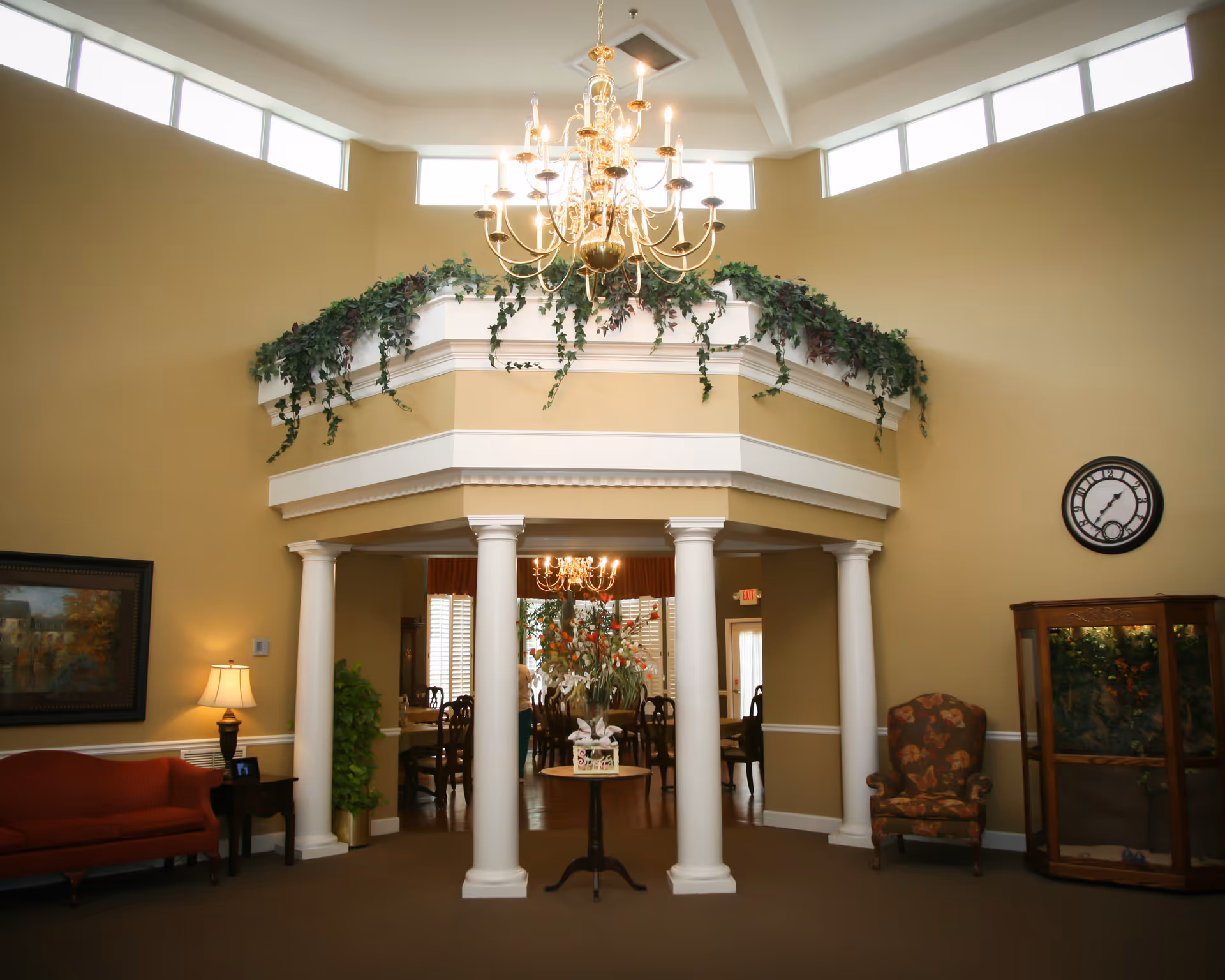 Bright, elegant senior living common area with a central chandelier over white columns, seating, and decorative greenery.
