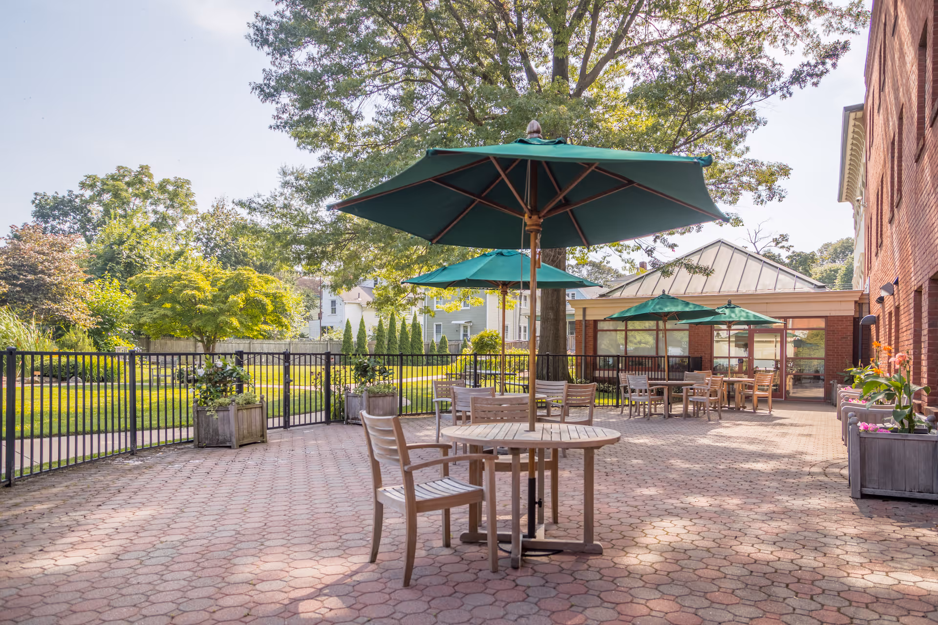 Outdoor patio with round wooden tables, chairs and green umbrellas next to a brick building and landscaped lawn.
