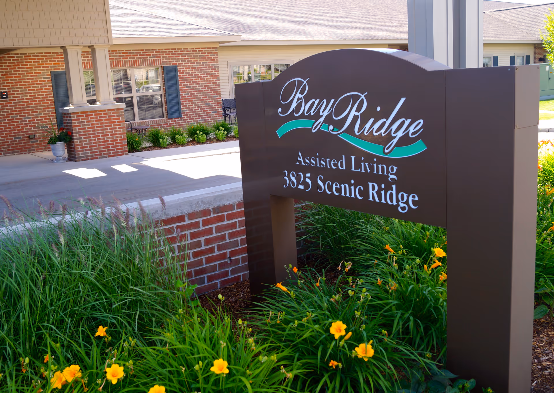 Outdoor view of the entrance area of Bay Ridge Assisted Living Center showing a brown sign with white and green lettering surrounded by green plants and yellow flowers, with a brick building and covered walkway in the background.