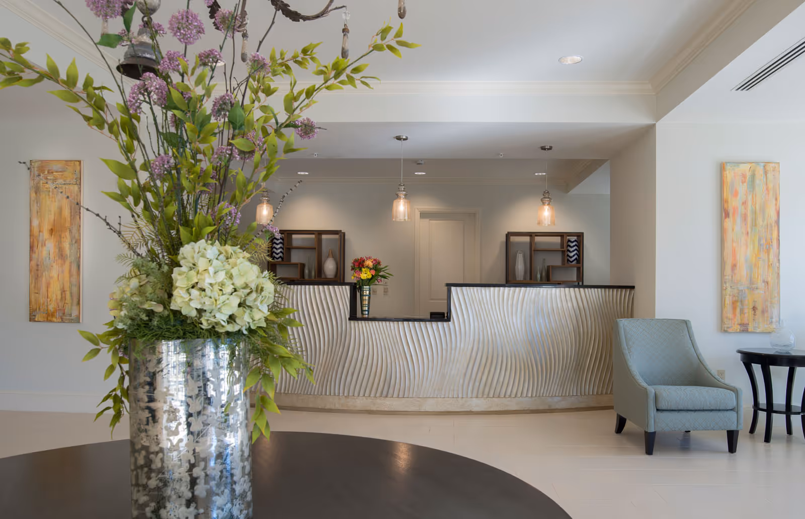 A modern reception area with a curved textured front desk, two pendant lights hanging from the ceiling, and decorative shelves behind the desk. In the foreground, there is a large silver vase with a floral arrangement featuring green leaves and purple and white flowers. To the right, there is a light blue upholstered armchair next to a small round black table. Two abstract paintings hang on the walls on either side of the reception desk.