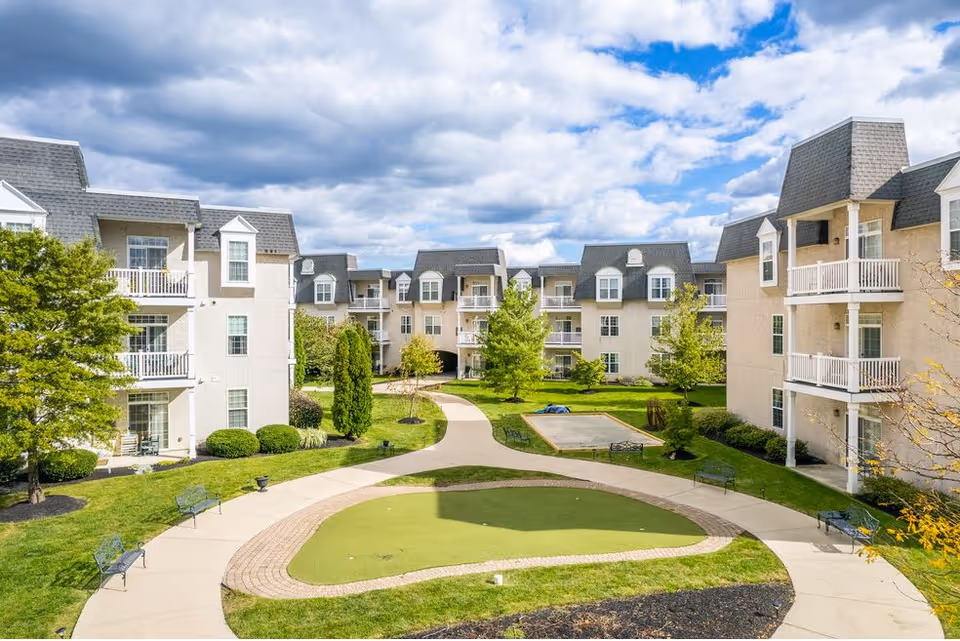 Outdoor courtyard area of a senior living facility with paved walkways, benches, green grass, trees, and multi-story buildings with balconies surrounding the space under a partly cloudy sky.