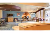 A spacious common area with multiple seating arrangements including chairs and tables. The room features a blue carpet, a flat-screen TV mounted on a wooden cabinet, and a decorative circular wall element with warm lighting. Large windows with curtains allow natural light to enter the space.