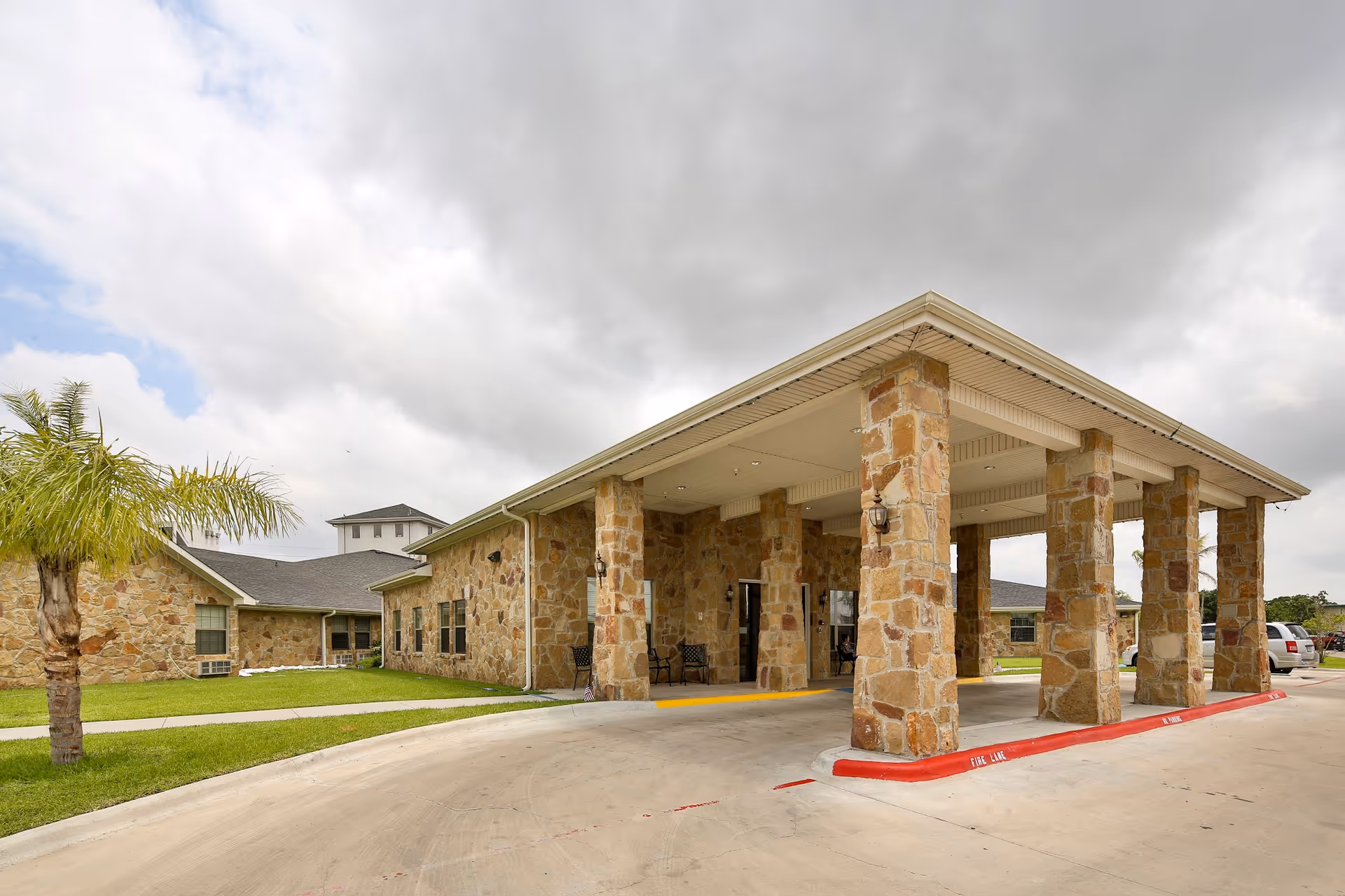 Exterior view of a nursing and rehabilitation center building with stone pillars supporting a covered entrance. The building has stone walls, a gray roof, and a driveway with a red fire lane curb. There is a small palm tree and green grass on the left side under a cloudy sky.