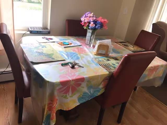A dining table covered with a colorful floral tablecloth, surrounded by four red cushioned chairs. On the table are placemats, a vase with pink and purple flowers, a set of keys, some papers, and a napkin holder. The room has wooden flooring and a window letting in natural light.