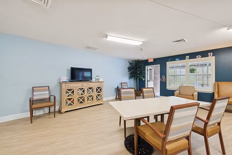 A bright and clean common area with light blue and dark blue walls, featuring a rectangular table surrounded by wooden chairs with cushioned seats. There is a wooden cabinet with a TV on top against one wall, additional chairs along the walls, a large window with white blinds, and a potted plant near an open door leading outside.