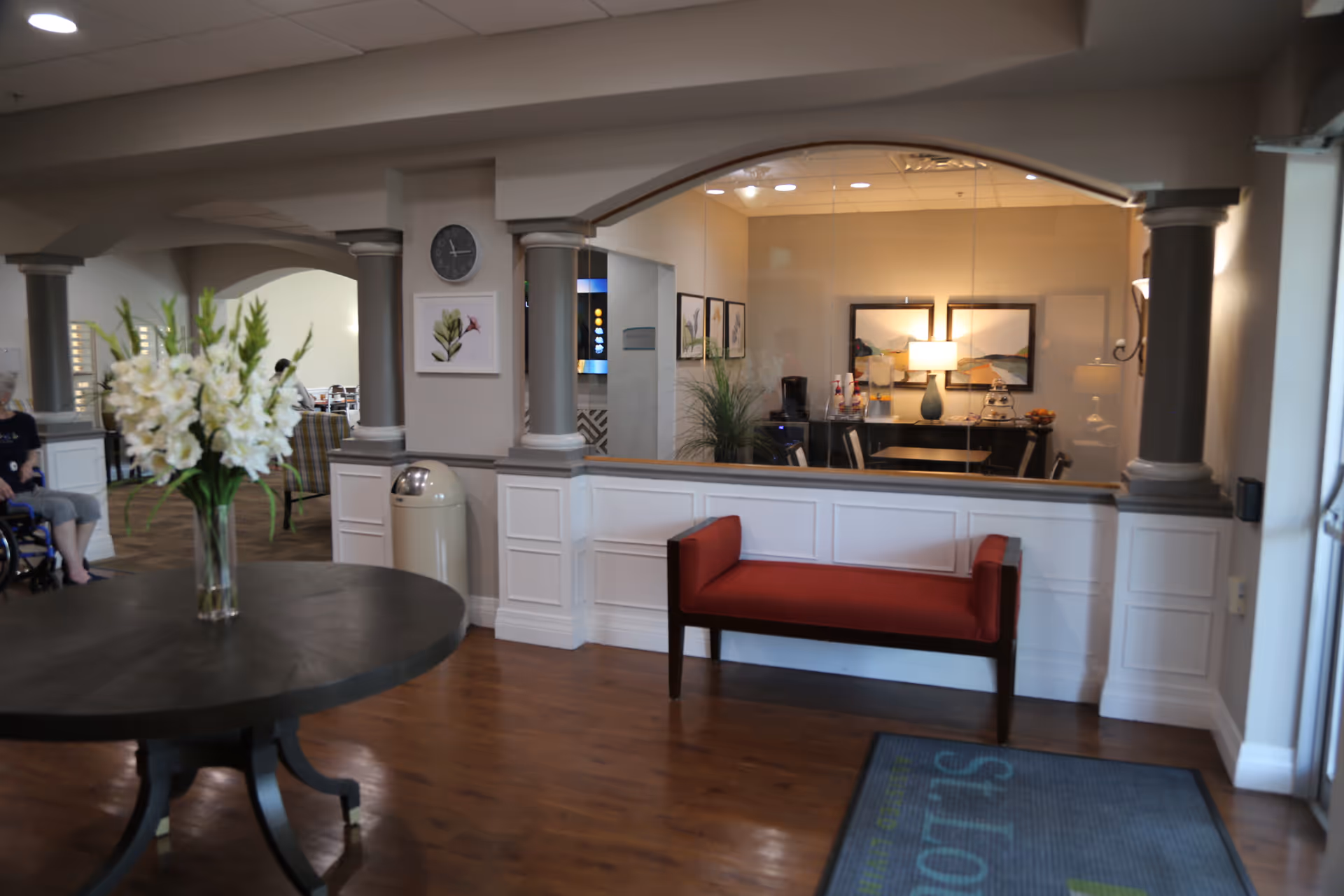 Interior view of a senior living facility lobby area with a round table holding a vase of white flowers, a red bench against a half wall with columns, and a reception or office area visible through a large window. There is a clock and framed artwork on the walls, and a person in a wheelchair is partially visible on the left side.
