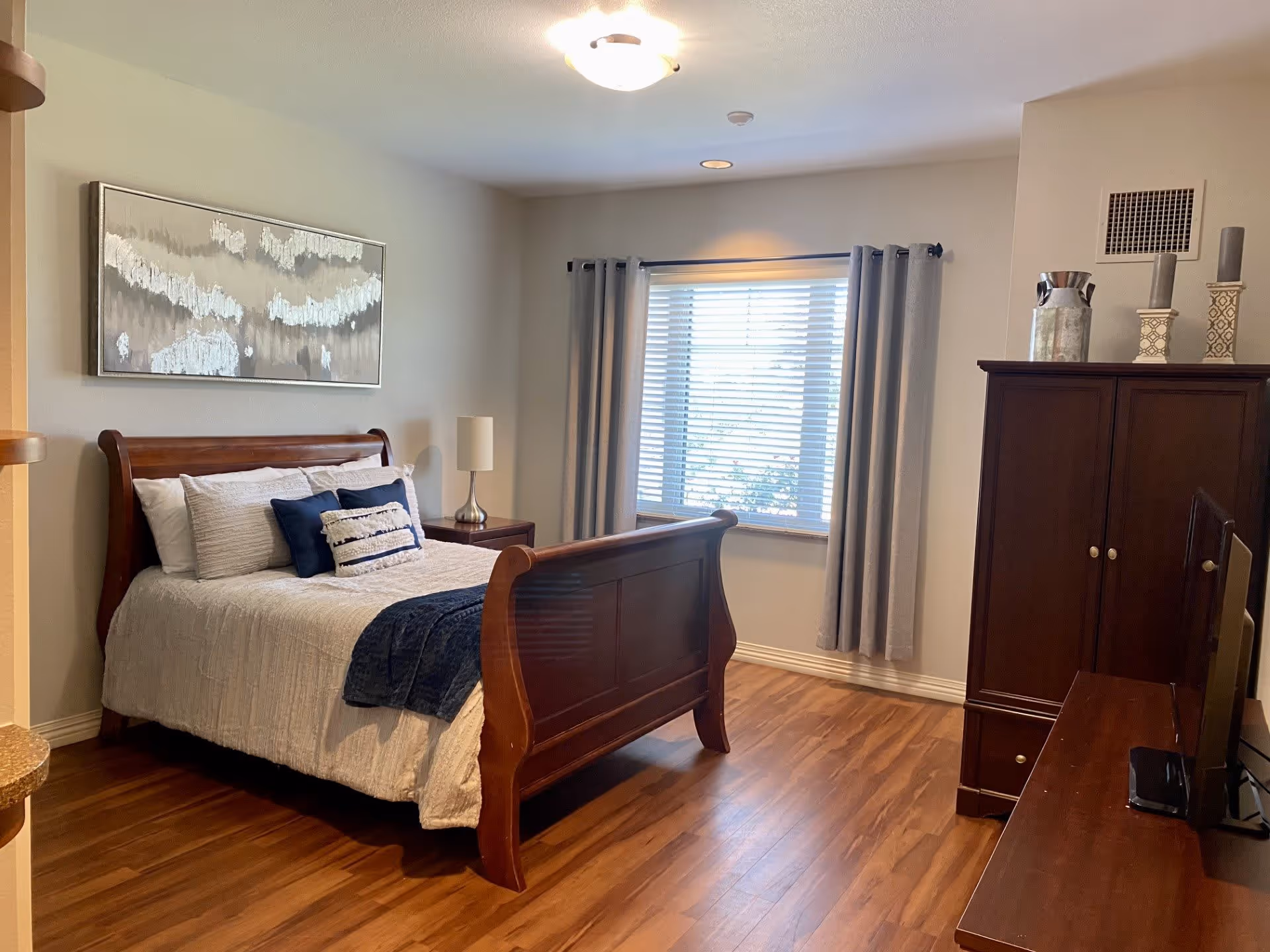 A bedroom with a wooden sleigh bed made with white and blue bedding, a nightstand with a lamp, a window with gray curtains, a wooden dresser with decorative items, and a TV on a wooden stand. The room has wood flooring and neutral-colored walls.