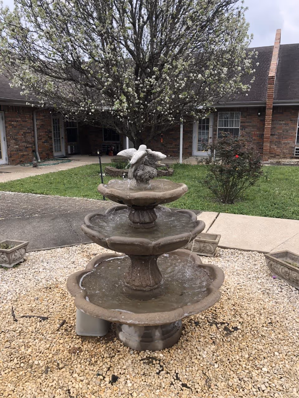 A three-tiered stone fountain in a gravel courtyard with a blooming tree and brick building entrances in the background.