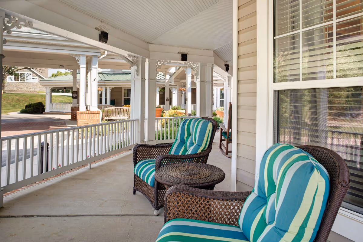 Covered outdoor porch area with two wicker chairs featuring green and blue striped cushions and a small round wicker table between them. The porch has white railings and columns with decorative trim, overlooking a paved driveway and landscaped garden area.