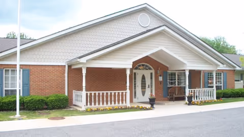 Front exterior view of a single-story brick building with a covered entrance supported by white columns, blue shutters on the windows, a bench on the porch, and neatly trimmed bushes and flowers along the walkway.