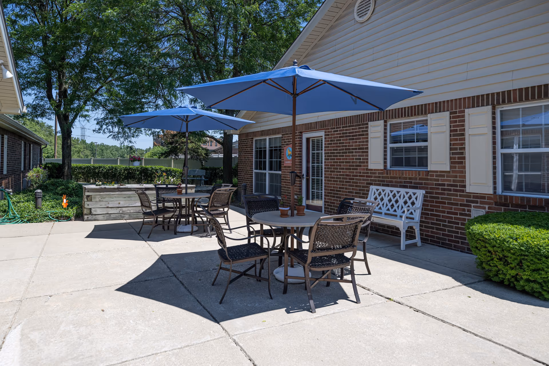 Outdoor patio area at Arden Courts - ProMedica Memory Care Community with round tables and chairs under blue umbrellas, a white bench against a brick wall, green bushes, trees, and a garden hose on the left side.
