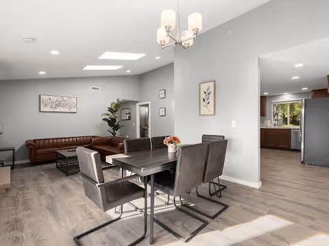 A modern open-concept living and dining area with a dark wood dining table and six upholstered chairs. The living room features a brown leather sectional sofa, a coffee table, and a large plant in the corner. The walls are painted light gray with framed artwork, and the flooring is wood. A kitchen area with stainless steel appliances is visible in the background.