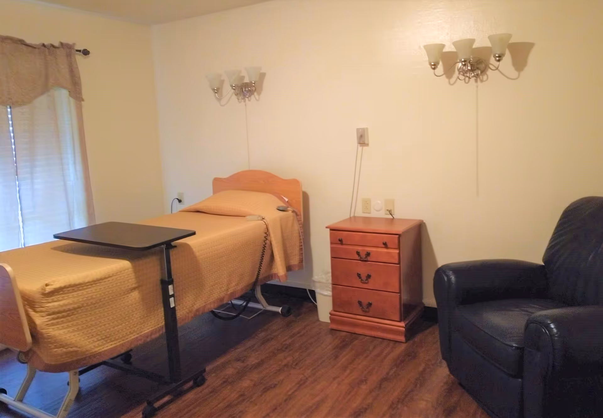 A nursing facility room with a single hospital-style bed covered with a beige blanket, a small wooden nightstand with drawers, a black leather armchair, and two wall-mounted light fixtures. There is a window with curtains on the left side and a wooden floor.
