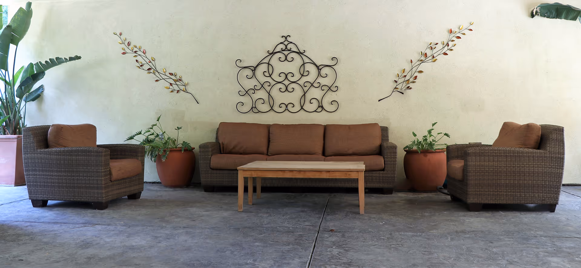 Wicker sofa and two matching armchairs with cushions arranged around a wooden coffee table on a covered patio with potted plants and wall decor.
