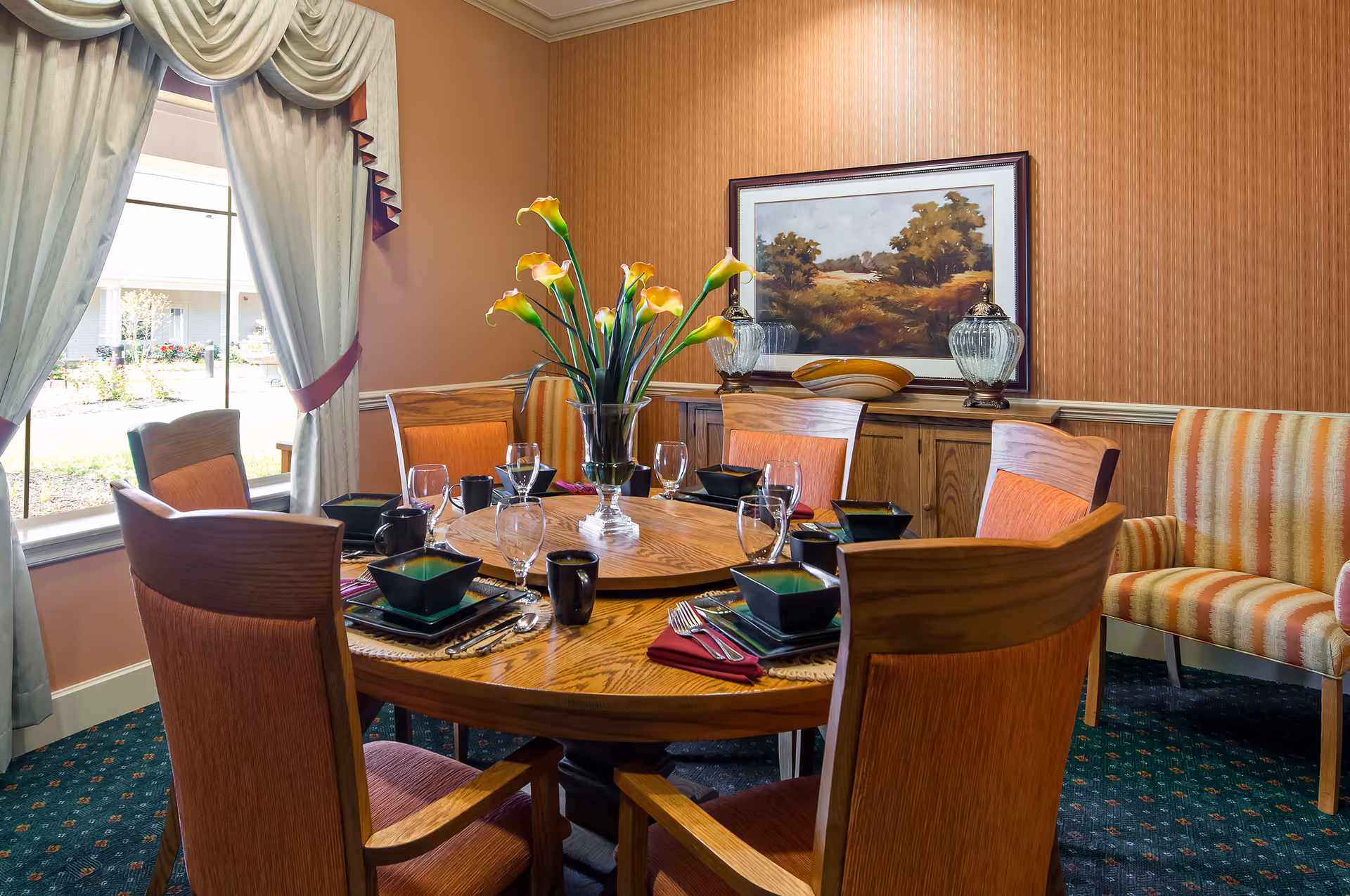 A dining room with a round wooden table set for six people. The table has black square bowls, black mugs, wine glasses, and silverware on red napkins. A vase with yellow calla lilies is in the center. The room has a large window with draped curtains, a wooden sideboard with decorative items, and a framed landscape painting on the wall. There is also a striped upholstered bench against the wall.