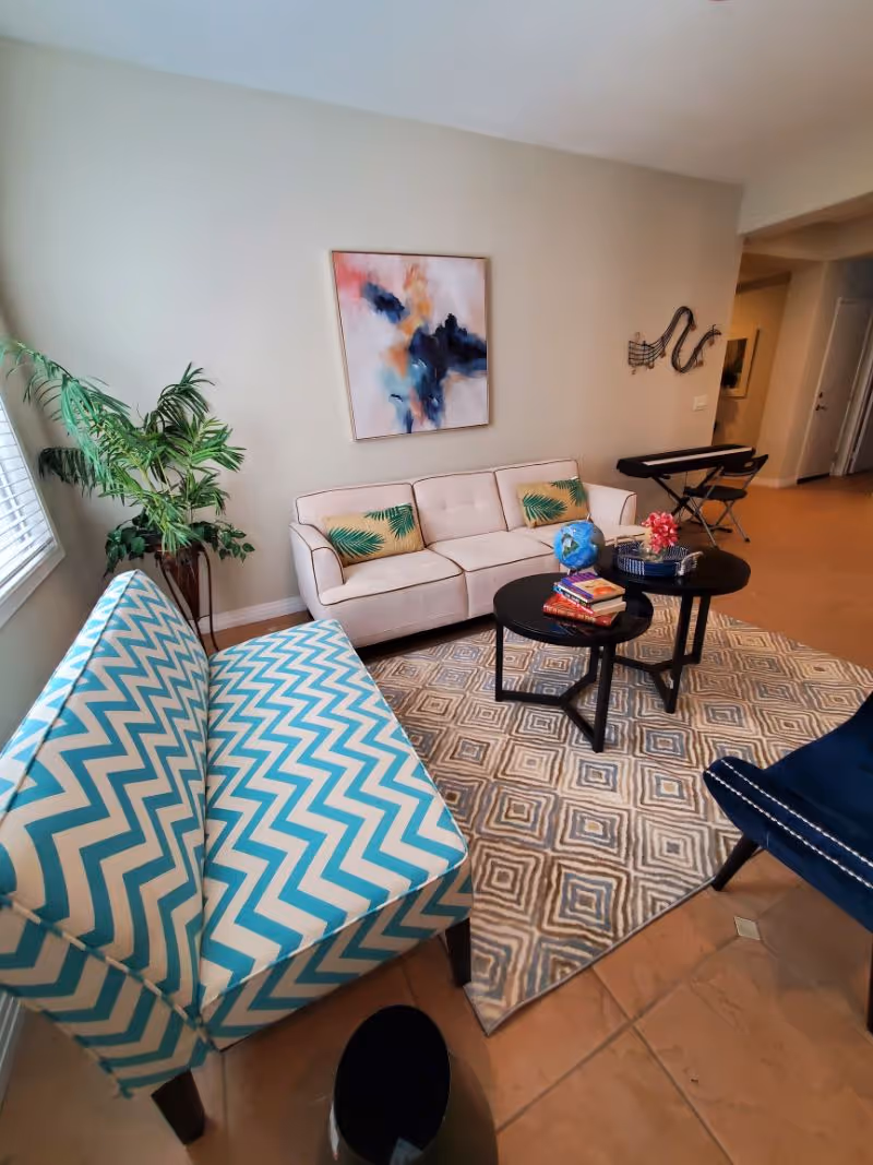 A cozy living room area with a white sofa adorned with green leaf-patterned pillows, a blue and white chevron-patterned bench, a blue armchair, and two black round coffee tables on a patterned rug. There is a colorful abstract painting on the wall, a potted plant near the window, and a keyboard with a chair in the background.