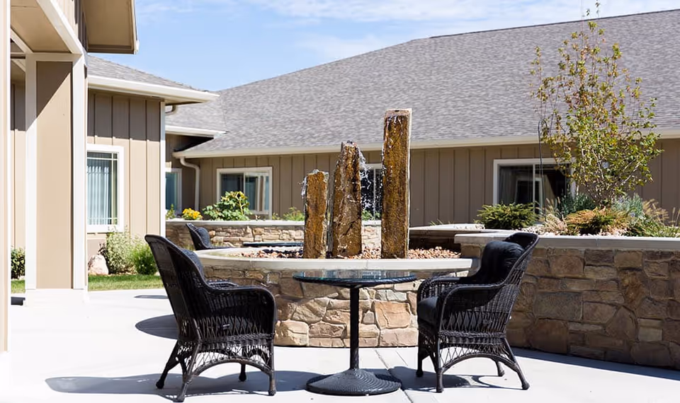 Outdoor courtyard showing a round table with wicker chairs and a stone water fountain in front of a single-story building.