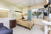 Bay Crest Care Center patient bedroom with single bed, sofa, dresser, sink, window, and blue curtains.