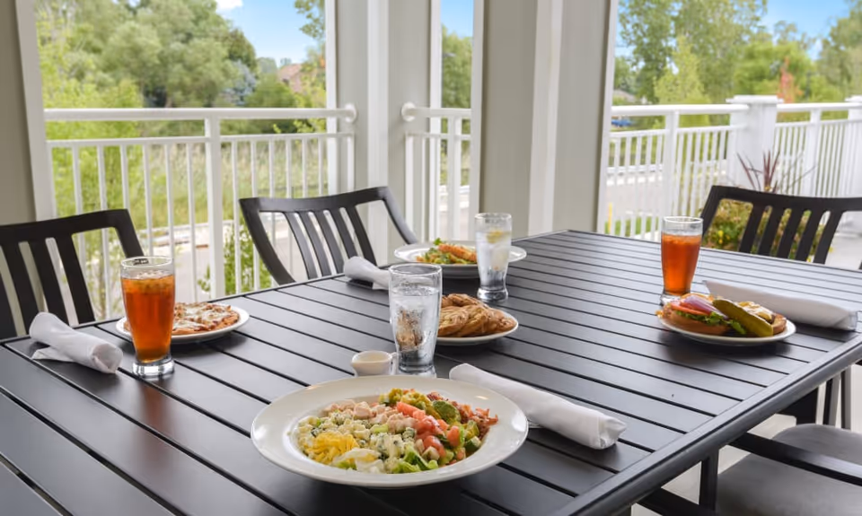 Outdoor dining table set with plates of food including a salad, sandwich, pizza, and pastries, along with glasses of iced tea and water. The table is on a patio with black chairs and white railings, surrounded by greenery.