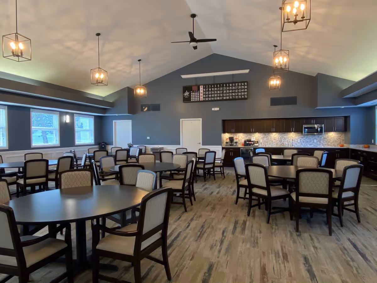 A spacious dining room with multiple round tables and cushioned chairs arranged neatly. The room features a high vaulted ceiling with modern pendant lights and ceiling fans. Along one wall, there is a kitchenette area with dark cabinets, a microwave, and a coffee maker. Large windows allow natural light to brighten the space.