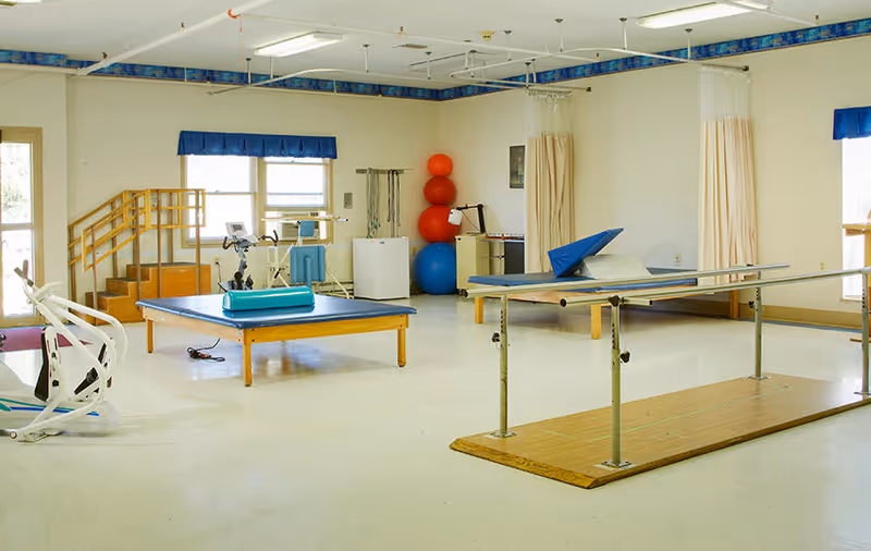 A spacious physical therapy or rehabilitation room with exercise equipment including parallel bars, therapy tables, exercise balls stacked in a corner, and a stationary bike. The room has large windows with blue valances and beige curtains, and a small wooden staircase with handrails.
