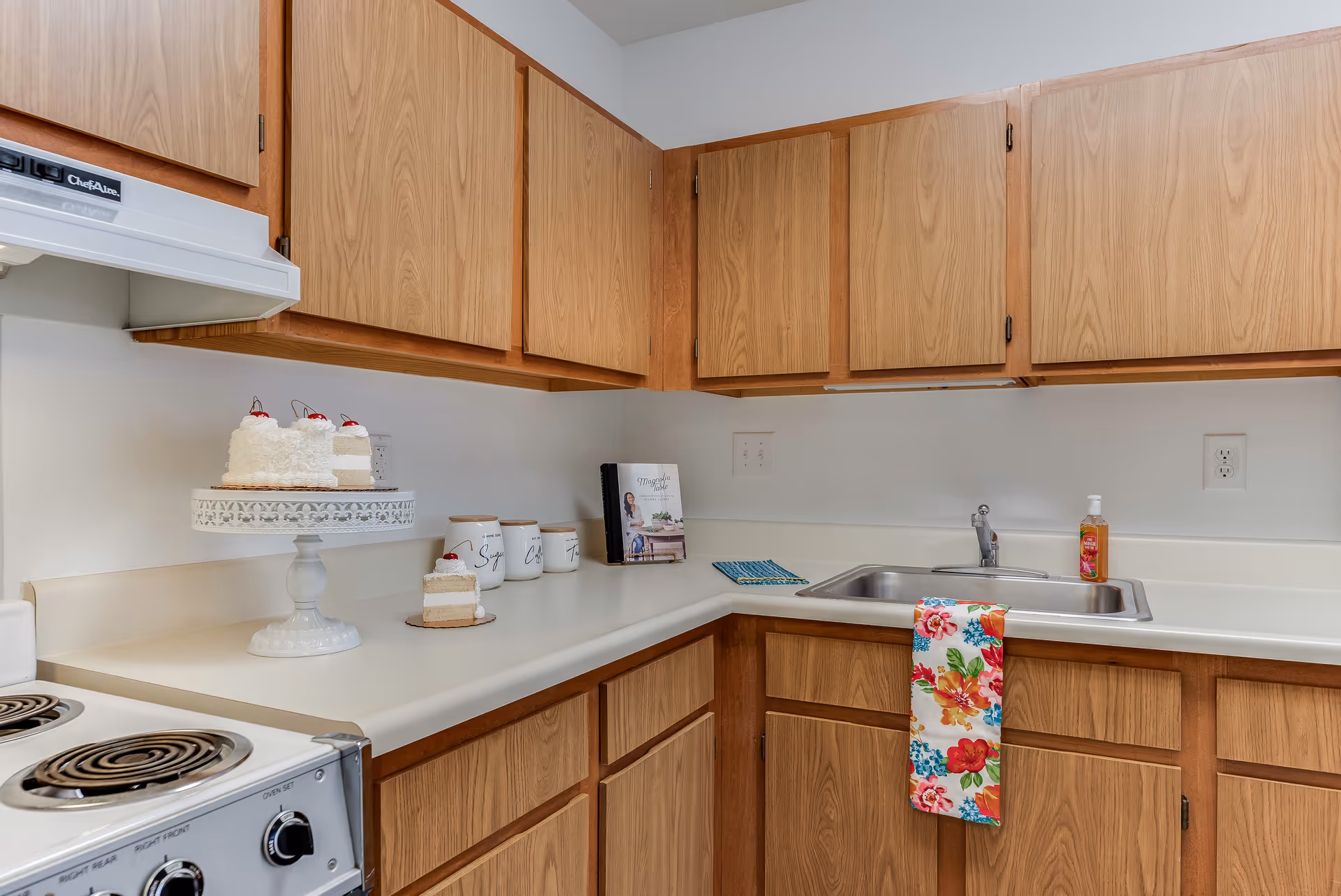 A kitchen corner with wooden cabinets above and below a white countertop. On the counter, there is a white cake stand holding a cake with white frosting and cherries on top, three white canisters labeled 'Sugar', 'Coffee', and 'Tea', a cookbook, a colorful floral dish towel hanging over the edge of the sink, and a bottle of hand soap next to the stainless steel sink. A white stove with coil burners is partially visible on the left side.