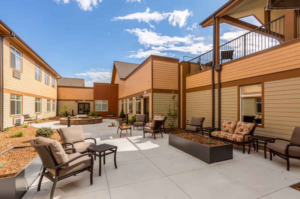 Outdoor courtyard area of Sierra Basin Memory Care facility with multiple cushioned chairs and small tables arranged on a concrete patio. The building exterior features beige and brown siding with windows, and there is a fire pit in the center surrounded by seating. The sky is blue with some clouds.