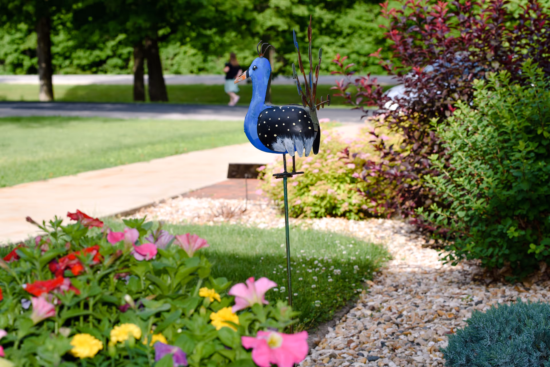 A colorful garden scene featuring a metal bird sculpture with a blue head and black body with white spots, standing on a metal stake among vibrant flowers and green bushes. A paved walkway and grassy area are visible in the background with trees and a person walking in the distance.