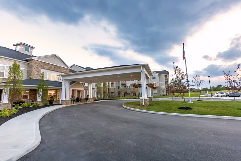 Covered porte-cochère and driveway leading to the front entrance of a senior living building with landscaped lawns and a flagpole.
