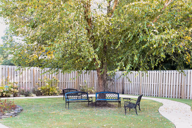 A peaceful outdoor garden area with a large tree at the center surrounded by four black metal benches arranged in a circle. The area is enclosed by a wooden fence, with a curved concrete pathway leading through the green lawn. There are some plants and flowers along the fence line.