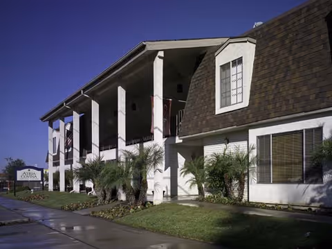 Exterior view of Atria Covina senior living facility showing a two-story building with white walls, large pillars, and a brown shingled roof. There are palm trees and well-maintained landscaping in front of the building under a clear blue sky.