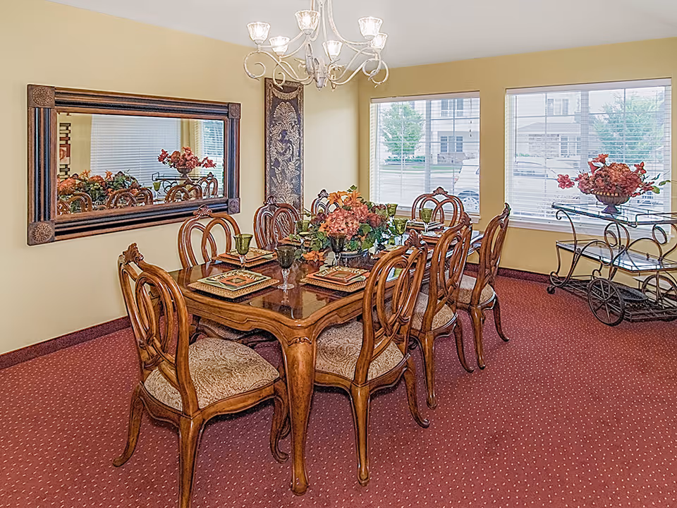 A formal dining room with a wooden dining table set for eight, featuring ornate chairs with cushioned seats. The table is decorated with floral centerpieces and place settings. The room has beige walls, a red carpet, a large rectangular mirror on one wall, three windows with blinds, and a chandelier hanging above the table. A decorative cart with flowers is positioned near the windows.