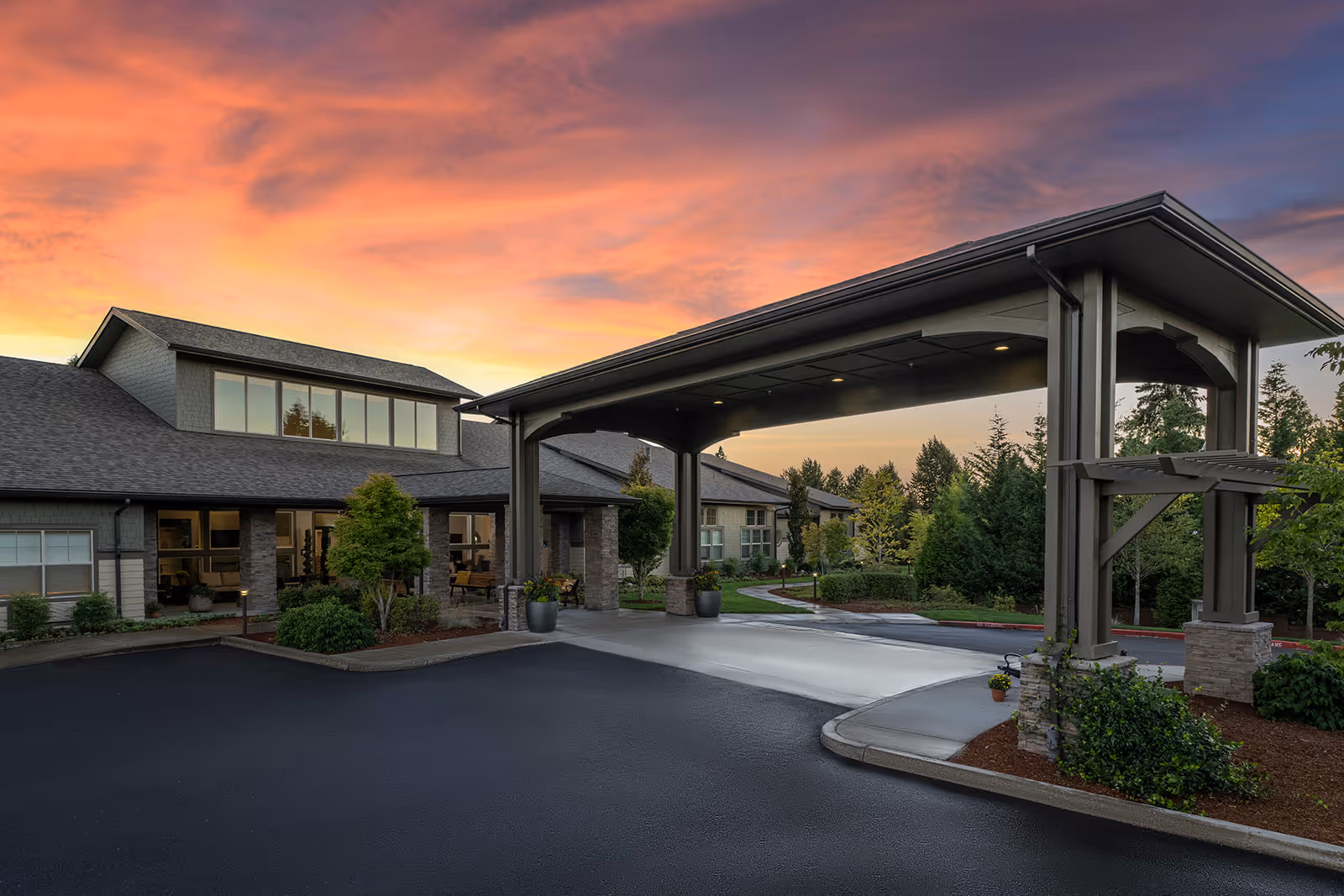 Exterior view of Waterhouse Ridge Memory Care facility at sunset, showing the main entrance with a covered drop-off area, landscaped greenery, and a clear sky with vibrant orange and purple hues.