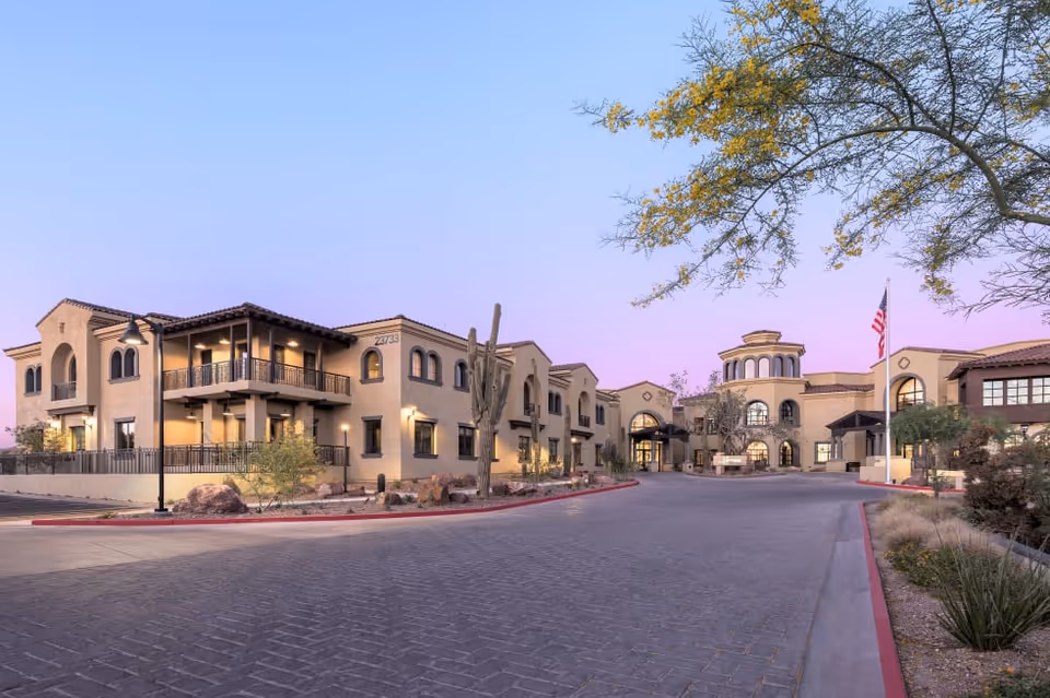 Exterior view of Clearwater Pinnacle Peak facility at dusk, showing a large, multi-story building with balconies, arched windows, desert landscaping including a tall cactus, and an American flag near the entrance.