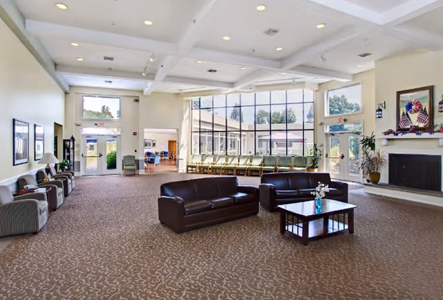 Spacious senior living facility common area with high ceilings, large windows letting in natural light, two dark leather sofas around a wooden coffee table with a flower vase, multiple chairs along the walls, and a fireplace decorated with flags and a wreath.