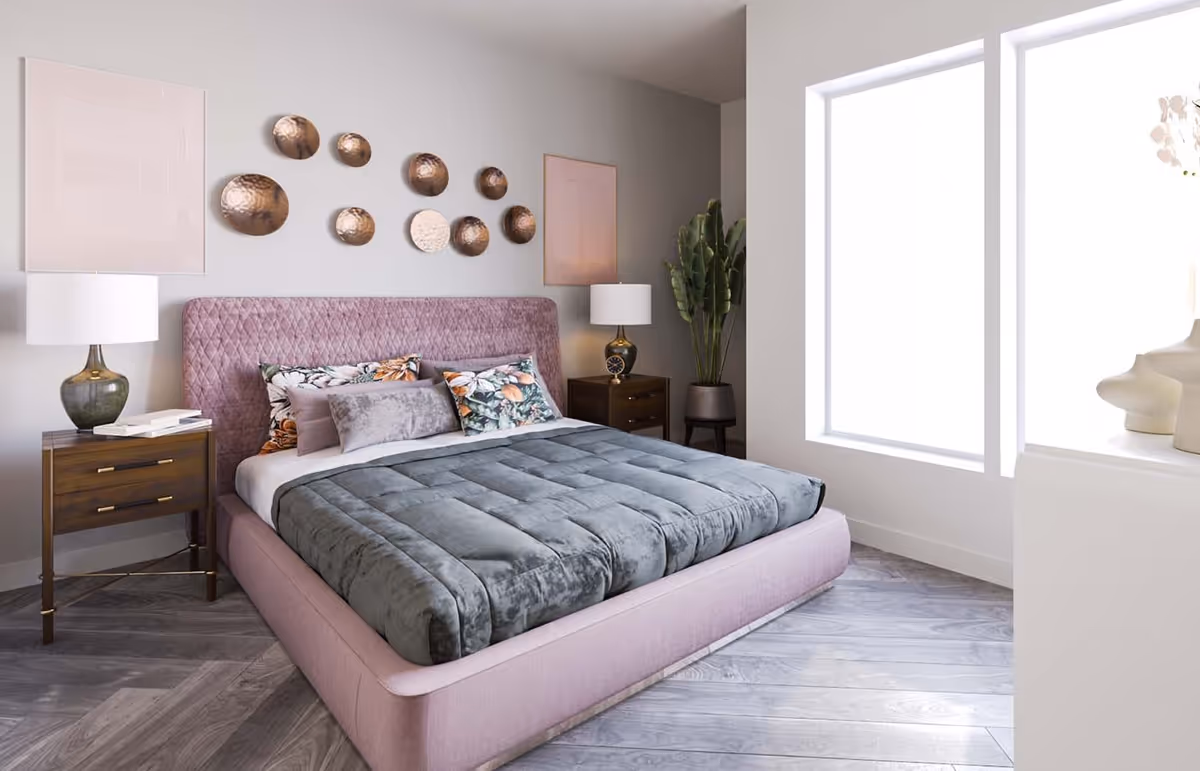 A modern bedroom with a pink upholstered bed frame and headboard, a gray comforter, and floral pillows. There are two wooden nightstands with lamps on either side of the bed. Decorative round metallic wall art is displayed above the headboard. A large plant is placed in the corner near two tall windows letting in natural light.