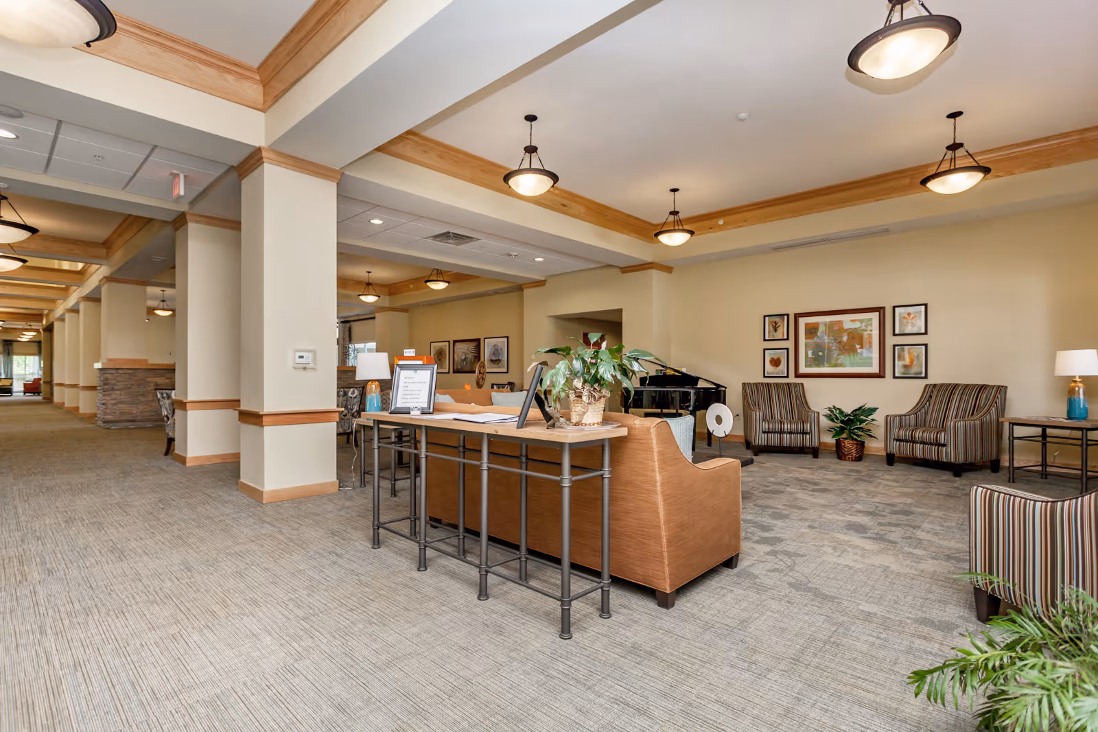 A spacious and well-lit senior living facility common area with beige walls and carpeted floors. The room features a seating area with striped armchairs, a brown sofa, a small table with a lamp, and framed artwork on the wall. There is a long hallway with columns and ceiling lights extending into the distance.